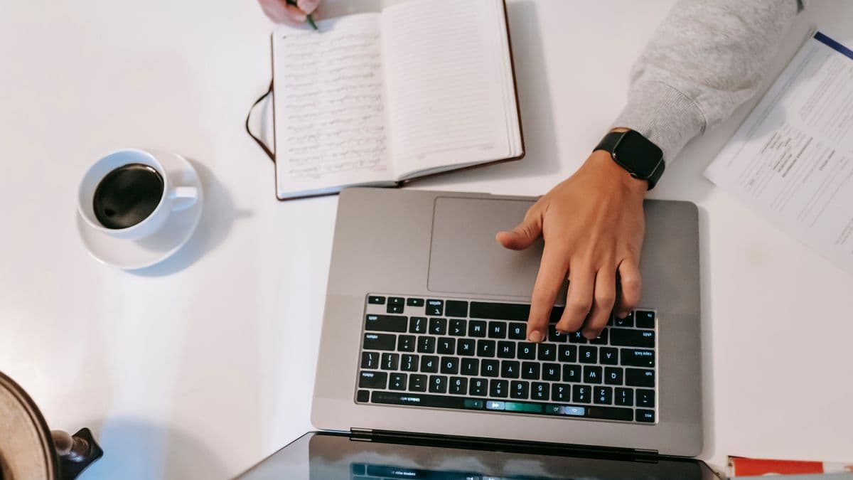 Manos escriben en una laptop junto a un cuaderno y un café